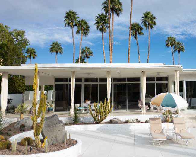 The image features a modern, single-story building with large glass windows, surrounded by palm trees and a landscaped patio area with lounge chairs and umbrellas.