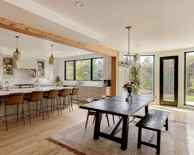 The image shows a modern open-concept kitchen and dining area with a large wooden table, high-backed chairs, and a view of the outdoors through large windows.