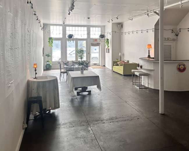 A spacious, well-lit interior of a minimalist café with tables and chairs arranged near large windows, and a counter in the background.