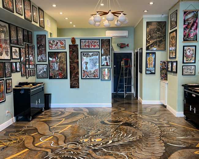 The image depicts a tattoo studio featuring a dragon-themed floor design, adorned with numerous framed artworks on the walls and various tools and equipment placed around the space.