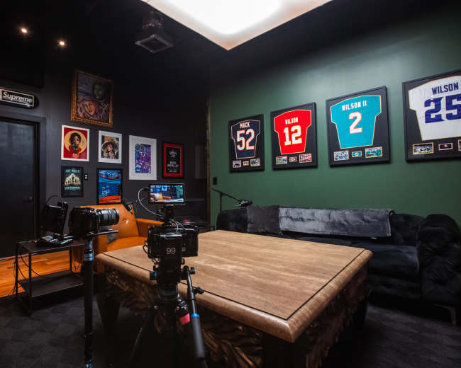 The image shows a dimly lit room with framed jerseys on the walls, a wooden table in the center, and cameras set up for recording.