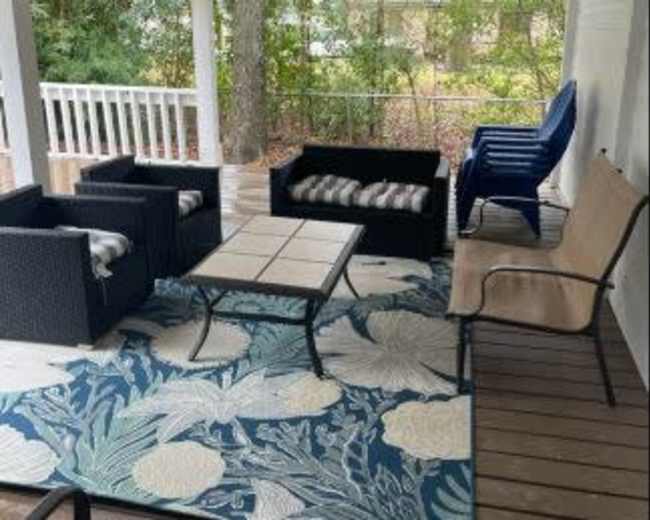 The image shows a covered porch area with various seating arrangements, including chairs and a table, situated on a patterned rug.