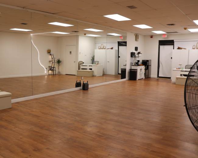 The image shows a spacious room with wooden flooring, a large mirror on one wall, and minimal furnishings including a bench and a small table.