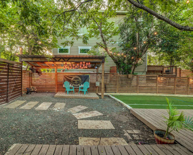A landscaped backyard features a wooden deck with two blue chairs, a pergola, and a pathway leading to a fenced area.