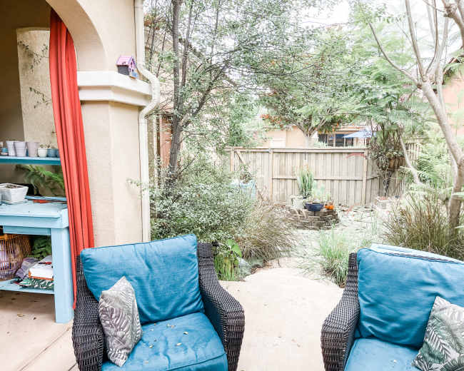 The image shows a small outdoor seating area with two blue chairs facing a garden path surrounded by greenery and a wooden fence.
