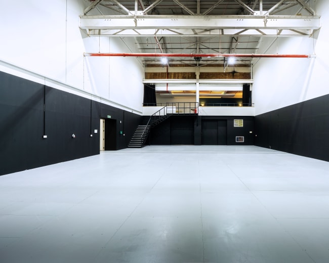 The image shows a large, empty industrial space with black walls and a white floor, featuring a staircase in the background.