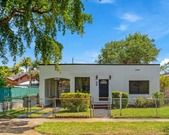 The image shows a single-story house with a dark front door, green landscaping, and a chain-link fence in a suburban setting.