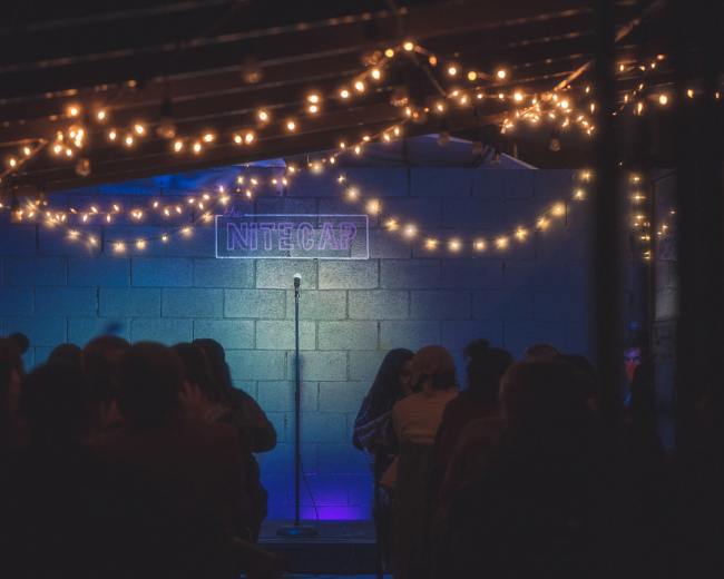 A dimly lit performance space features a microphone on stage, surrounded by string lights, with an audience seated in front.