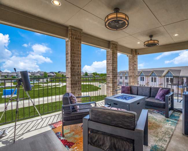 The image shows a balcony setup with outdoor furniture, including a seating area and a fire pit, overlooking a green space and residential buildings.