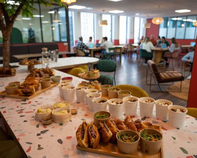 A variety of food items are displayed on a table in an office setting, with people working in the background.