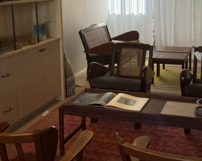 The image shows a cozy interior with wooden furniture, shelving containing decorative items, and natural light filtering through sheer curtains.