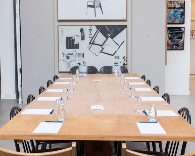 A long wooden conference table is set with water bottles and notepads, surrounded by black chairs in a room featuring framed artwork on the walls.