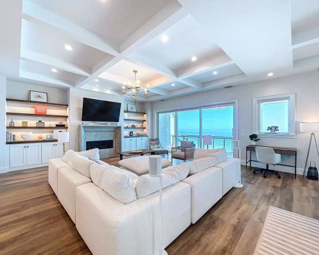 A spacious living room features a large sectional sofa, a flat-screen TV mounted on the wall, and a desk by a window with a view of the ocean.