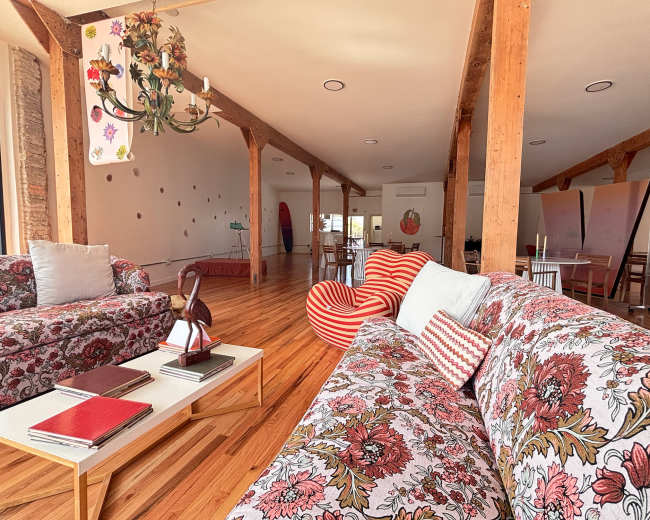 The image shows a spacious, brightly lit room with wooden beams, floral-patterned sofas, and various seating arrangements, including a striped chair and surfboards in the background.
