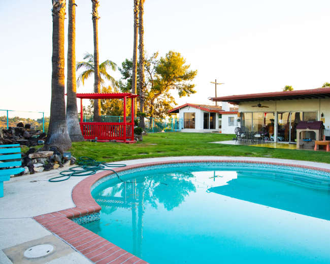 The image shows a backyard scene featuring a swimming pool, a red playground structure, and a house with a patio area surrounded by palm trees.