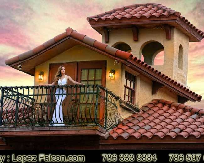 A woman stands on a balcony with a wrought iron railing on a house featuring a tiled roof against a vibrant sunset sky.