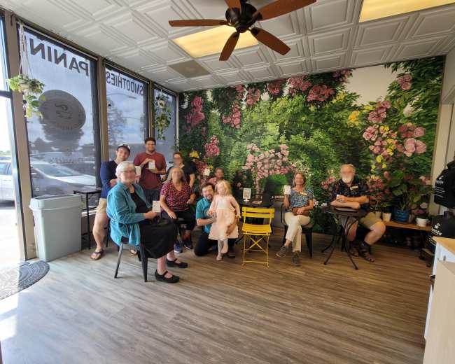 A group of people sits in a cafe with a floral mural on the wall and various plants around the space.