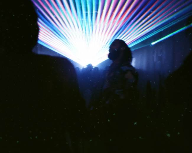 The image shows silhouettes of people in a dimly lit space with colorful laser lights projected across the walls.