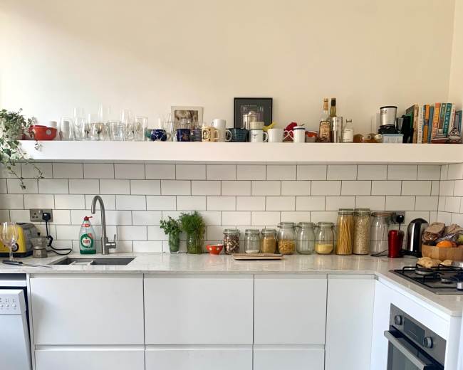 The image shows a modern kitchen with white cabinetry, a tiled backsplash, and various jars and utensils arranged neatly on open shelves.