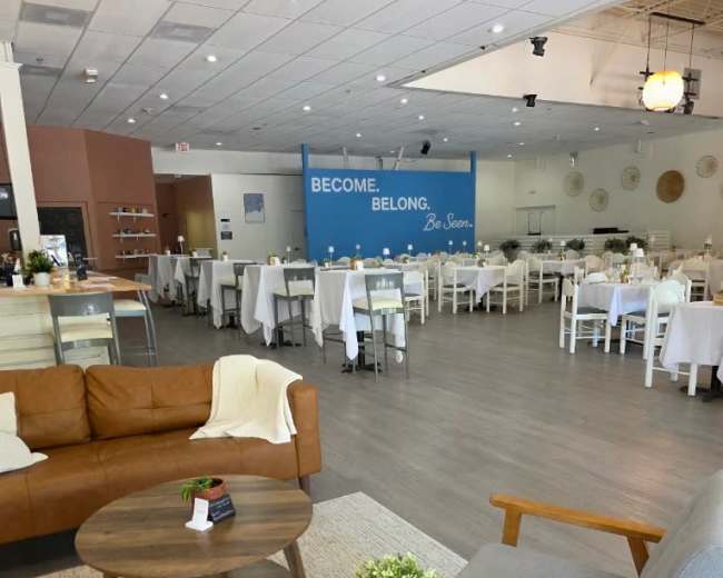 The image shows a modern, spacious restaurant interior with white tablecloths, wooden furniture, and a wall that has the text "BECOME. BELONG. Be Seen." prominently displayed.