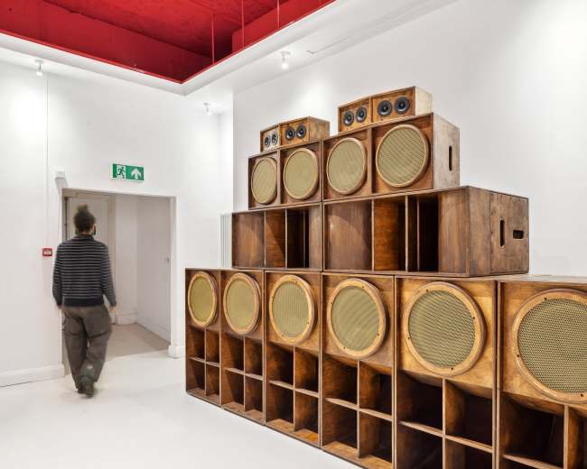 A person walks past a large set of stacked wooden speakers in a minimalist room with white walls and a red ceiling.