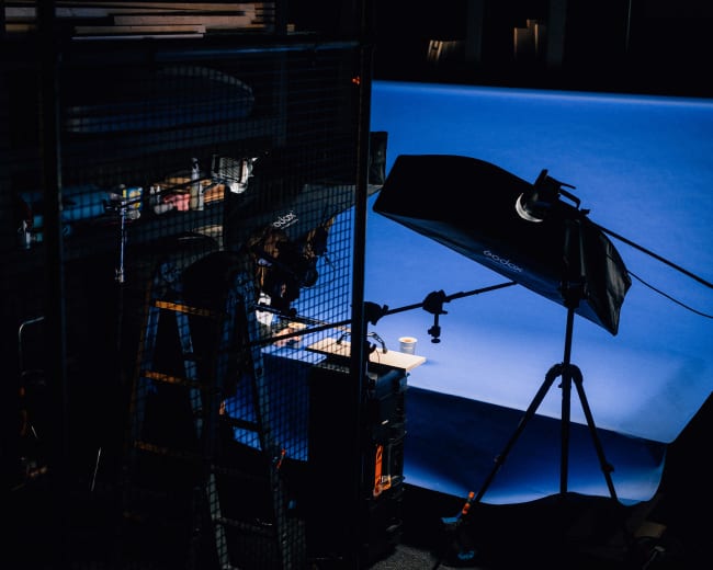 A photographer is positioned beside a ladder, adjusting equipment in a studio with a blue backdrop and various lighting setups.