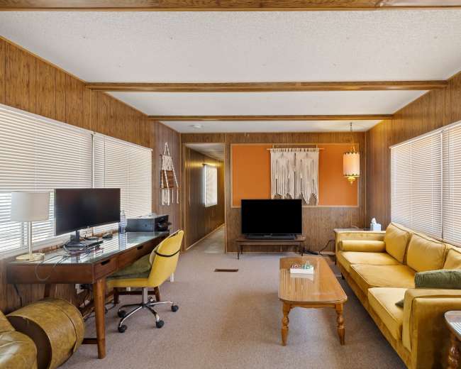 The image shows a cozy living room featuring a yellow sofa, a wooden coffee table, and a desk with a computer across from a television.