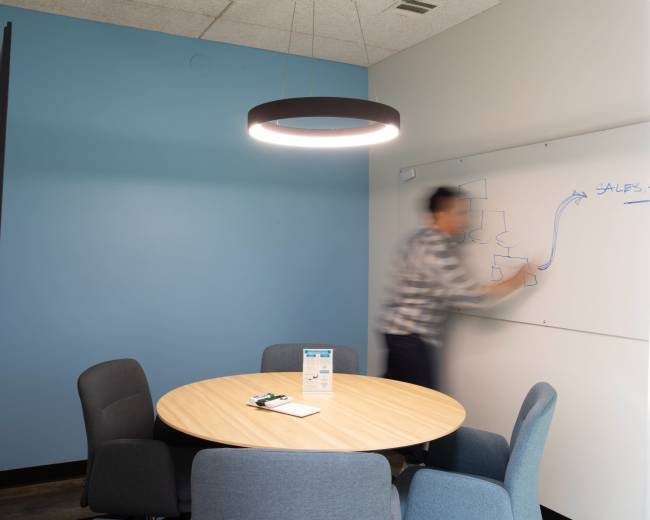 A person is writing on a whiteboard in a small conference room with a circular table and a mounted TV.