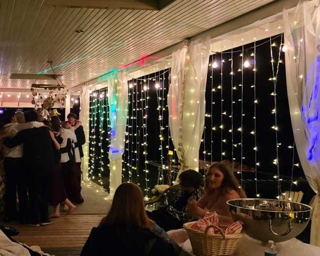 A group of people gather and dance on a lit porch adorned with fairy lights and decorations.