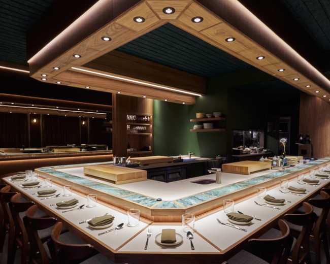 The image showcases a modern restaurant interior featuring a large sushi bar with an illuminated wooden canopy and a row of dining chairs arranged around a central countertop.