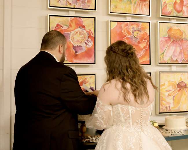 A couple stands together, with the man in a black suit and the woman in a lace wedding dress, facing a wall adorned with colorful floral paintings.