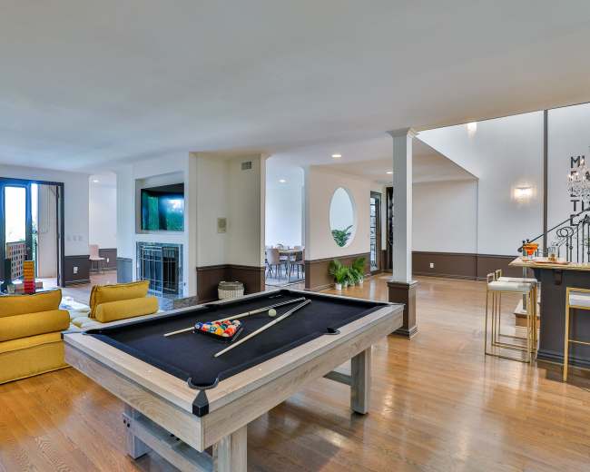 The image shows a spacious room featuring a pool table at the center, with a seating area and a dining space visible in the background.