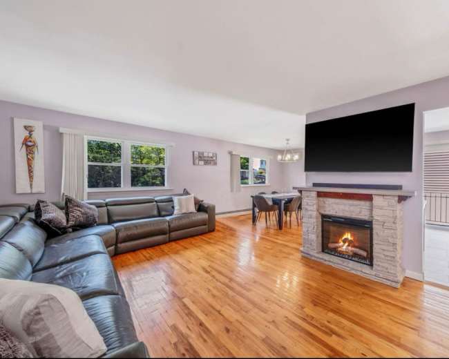 The image shows a spacious living room featuring a large sectional sofa, a stone fireplace, and a dining area with a table, illuminated by natural light from multiple windows.