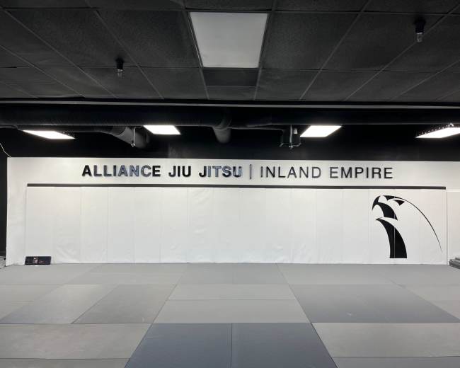 The image shows a jiu-jitsu training facility with a white wall featuring the text "ALLIANCE JIU JITSU | INLAND EMPIRE" and a graphic of a lion.