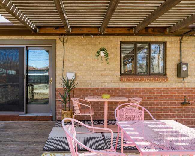 The image shows a patio area with a pink metal table and chairs, a potted plant, and sliding glass doors leading into a house.