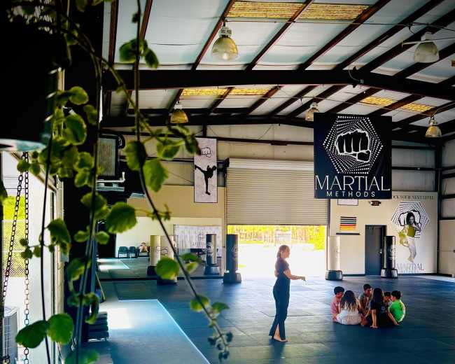 A group of children sits on a blue mat in a spacious martial arts studio, while an instructor stands in front of them.