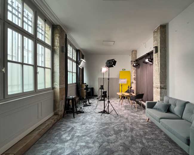 The image shows a well-lit studio space featuring a gray sofa, a wooden table with chairs, and professional lighting equipment set up against a backdrop.