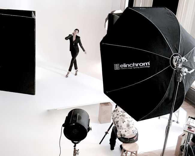 A model poses on a white backdrop during a photoshoot, while a large softbox and studio lights illuminate the scene.