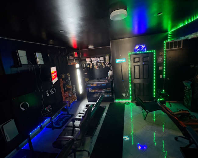 The image shows a dimly lit room with black walls, colorful lights, and multiple seating areas, likely designed for gaming or entertainment activities.