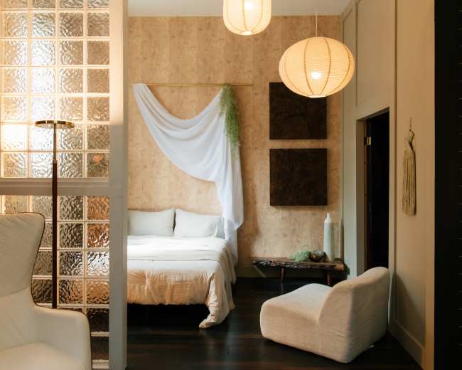 The image shows a softly lit bedroom featuring a bed with white linens, decorative wall hangings, and two round pendant lamps.