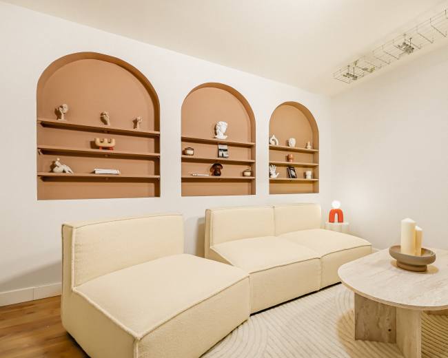 The image shows a modern, minimalist living room with two curvy sofas facing a round coffee table and threearched shelves displaying various sculptures on the wall.