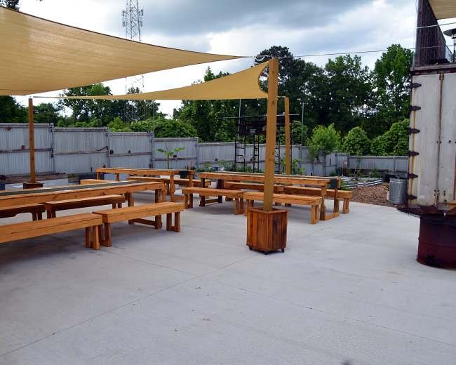 The image shows an outdoor seating area with wooden benches and tables shaded by large fabric canopies, surrounded by greenery and a gray fence.