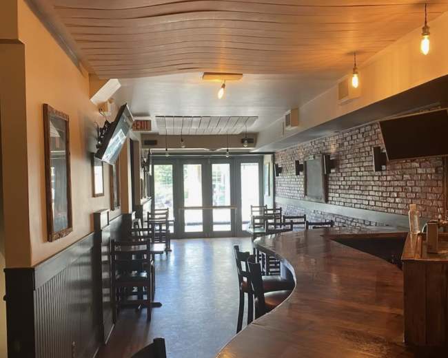 The image shows the interior of a restaurant or bar, featuring a curved wooden counter, empty tables and chairs, and large windows allowing natural light.