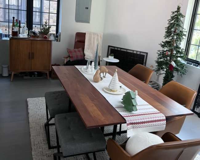 A dining room features a wooden table surrounded by chairs, decorated with a holiday table runner, while a Christmas tree stands in the corner.