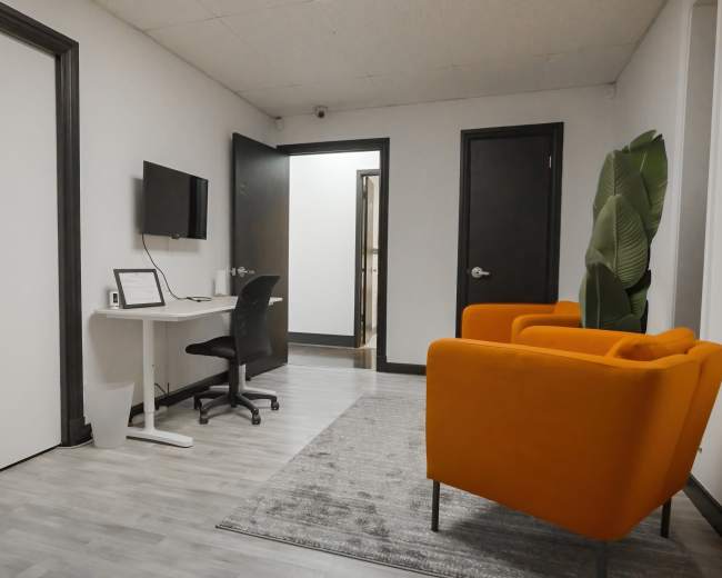 A modern office space featuring a desk with a computer, a black chair, an orange armchair, and a large plant, all set against a white wall and gray flooring.