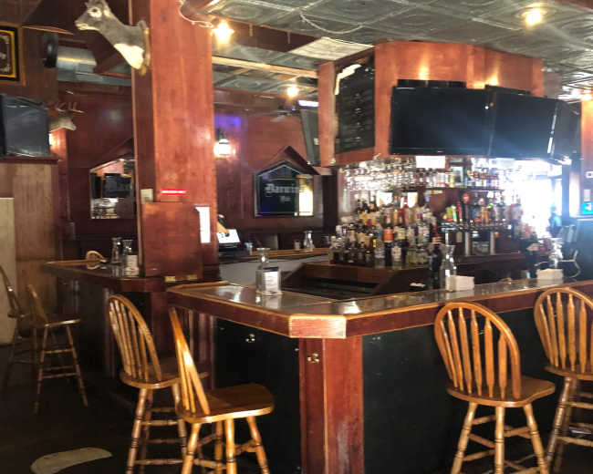 The image shows an empty bar area with a wooden counter, a variety of liquor bottles on display, and several wooden chairs arranged around it.