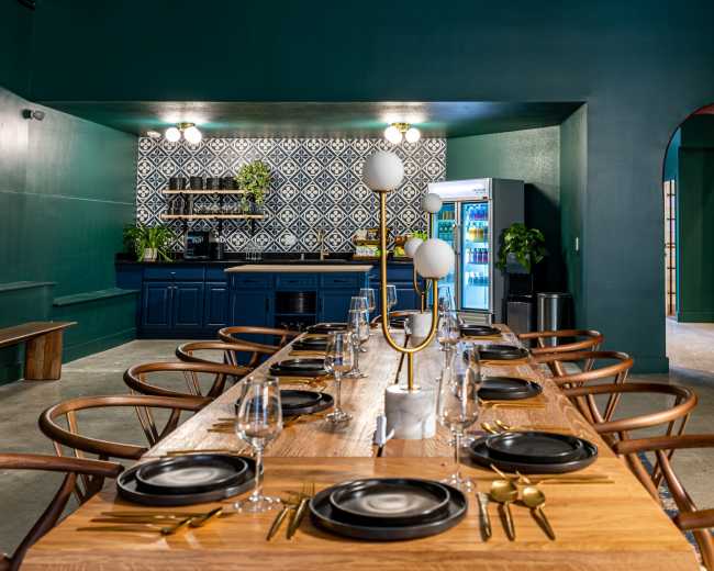 A modern dining area features a long wooden table set with plates and glassware, surrounded by stylish chairs, with a blue kitchen area and patterned backsplash in the background.