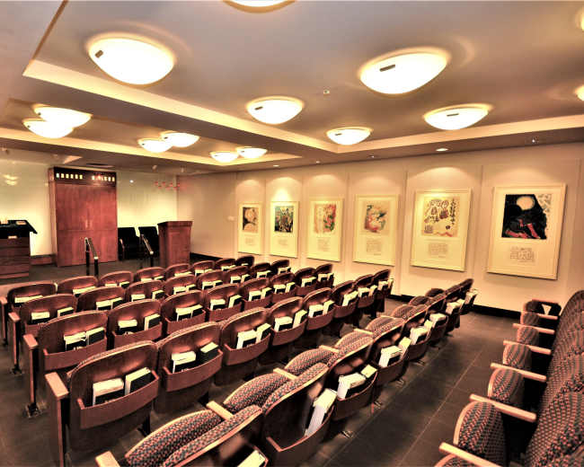 The image shows a conference room with rows of wooden chairs facing a podium, flanked by framed artworks on the walls.