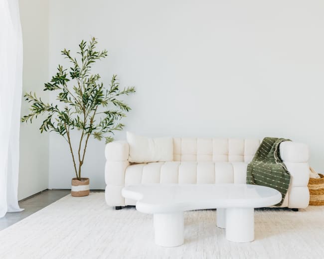 The image shows a modern living room featuring a white tufted sofa with a green throw, a round coffee table, and a potted plant.
