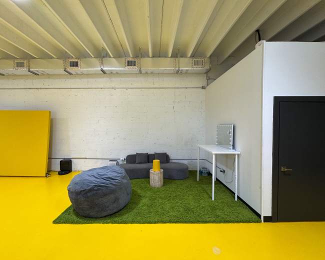 The image shows a spacious room with yellow flooring, a gray sectional sofa, a large bean bag, a small yellow table, and a mirror on a white wall.